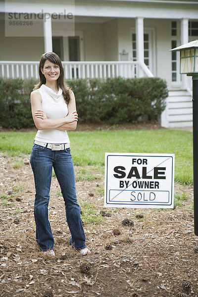 Eine junge Frau steht neben einem verkauften Schild vor einem Haus.