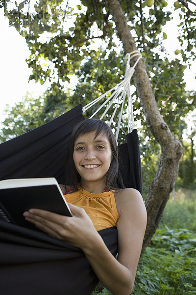 Junge Frau lächelt und liest Buch in Hängematte