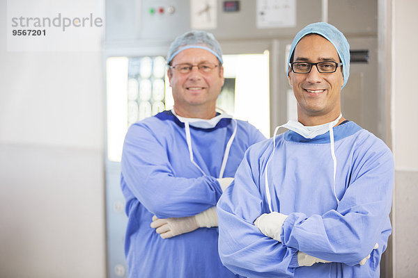 Zwei lächelnde Ärzte mit verschränkten Armen  in OP-Kleidung und Brille im Krankenhaus.