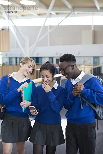 Drei glückliche Schüler in Schuluniformen mit Smartphone im Schulflur