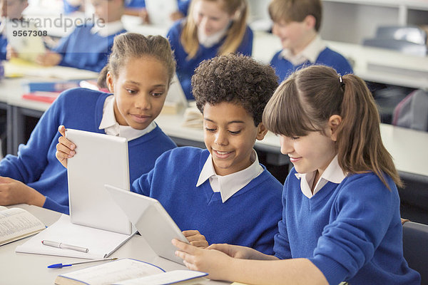 Grundschulkinder in blauen Schuluniformen mit digitalen Tabletts am Schreibtisch im Klassenzimmer