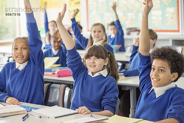 Grundschulkinder in blauen Schuluniformen  die im Klassenzimmer die Hände heben.