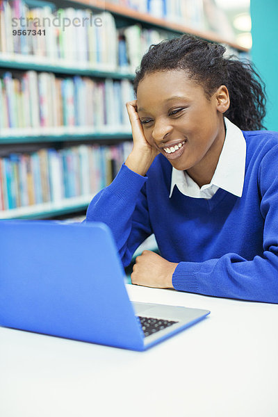 Lächelnde Studentin beim Blick auf den Laptop in der Bibliothek