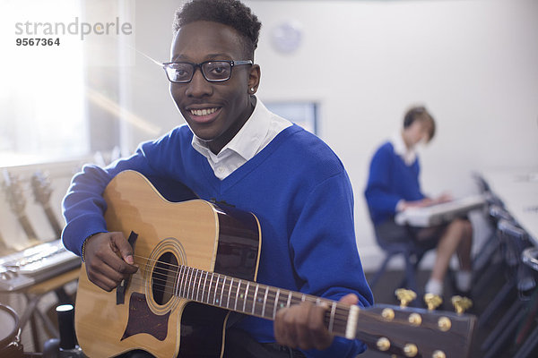 Porträt eines lächelnden Schülers  der im Klassenzimmer Akustikgitarre spielt.