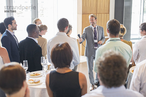 Porträt eines lächelnden Mannes  der im Konferenzraum steht und dem Publikum ein Mikrofon anbietet.