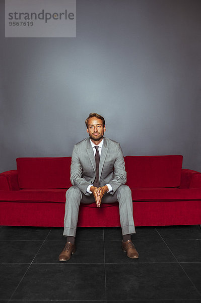 Porträt eines Geschäftsmannes im grauen Anzug sitzend mit den Händen in einem dunklen Raum.