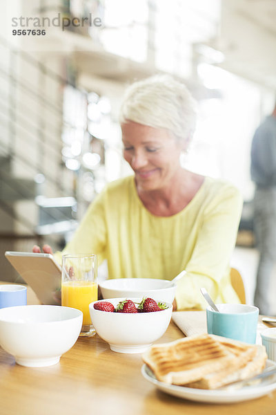 Ältere Frau mit digitalem Tablett am Frühstückstisch