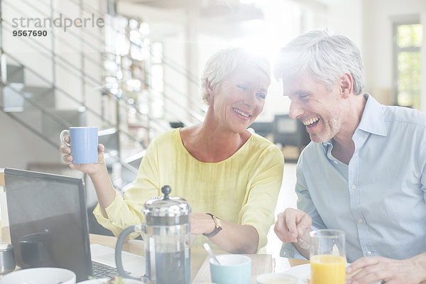 Älteres Paar lacht gemeinsam am Frühstückstisch mit Laptop