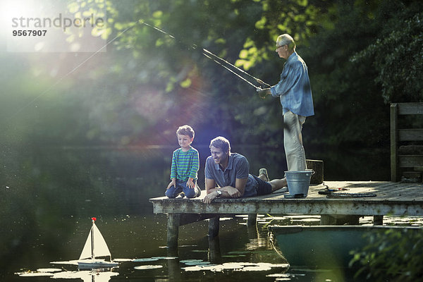 Junge fischen und spielen mit Spielzeug-Segelboot mit Vater und Großvater am See