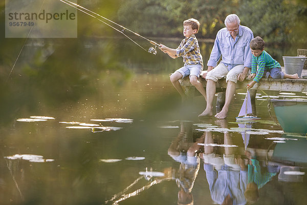 Großvater und Enkel fischen und spielen mit Spielzeug-Segelboot am See
