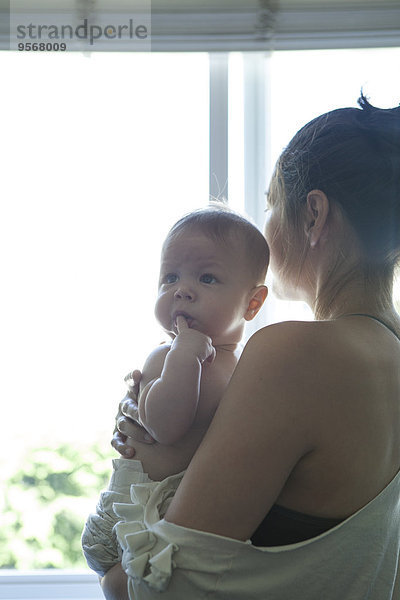 Mutter hält kleines Baby und schaut durchs Fenster.