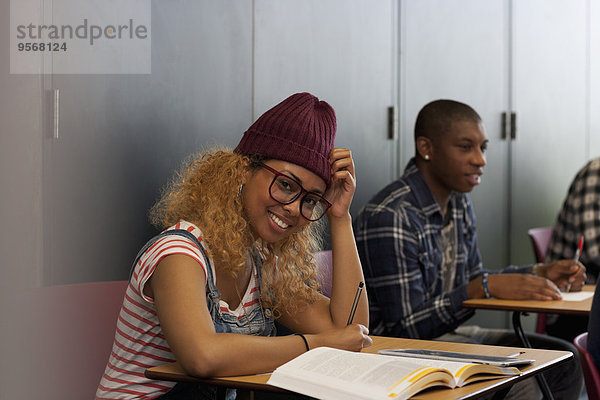 Studentin sitzt während der Vorlesung am Schreibtisch und lächelt vor der Kamera.