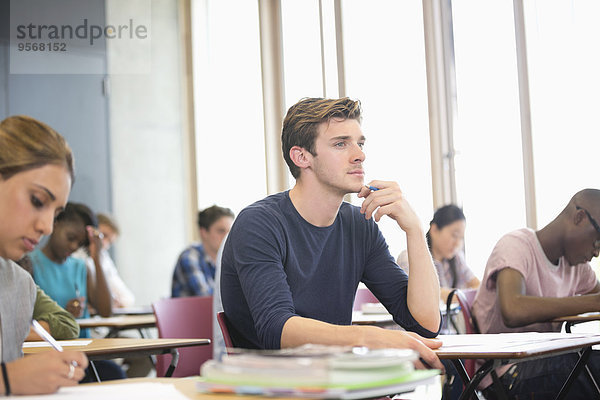 Männlicher Student mit Hand am Kinn während der Vorlesung mit anderen Studenten im Hintergrund