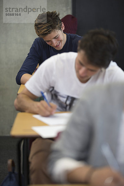 Blick auf lächelnde Schüler  die während einer Prüfung im Klassenzimmer am Schreibtisch sitzen