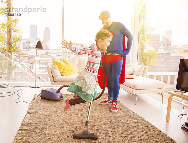 Superhelden-Vater beim Staubsaugen  während die Tochter im Wohnzimmer springt.