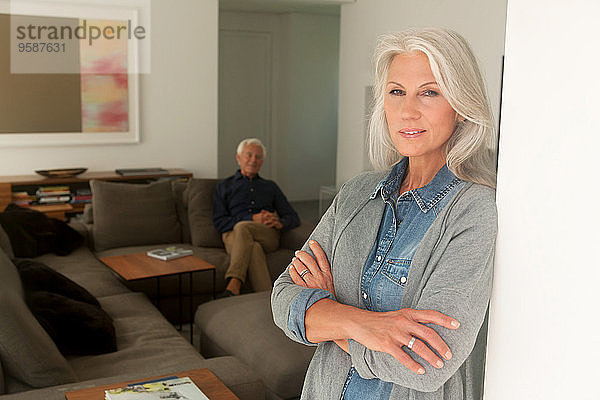 Porträt einer älteren Frau  die sich an eine Wand lehnt  während ihr Mann im Hintergrund auf der Couch sitzt.