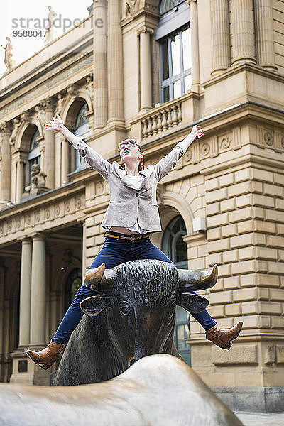 Deutschland  Hessen  Frankfurt  Geschäftsfrau mit ausgestreckten Armen auf Stierskulptur vor der Börse sitzend