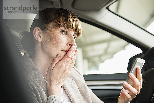 Porträt einer Geschäftsfrau  die in einem Auto sitzt und ihr Smartphone mit der Hand auf dem Mund betrachtet.