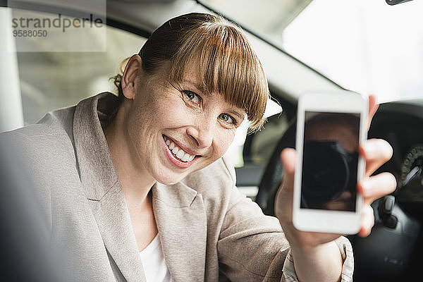 Porträt einer lächelnden Geschäftsfrau  die in einem Auto sitzt und ihr Smartphone zeigt.