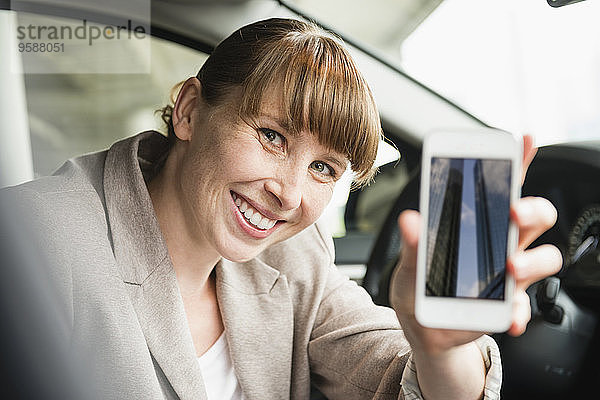 Porträt einer lächelnden Geschäftsfrau  die in einem Auto sitzt und ihr Smartphone zeigt.