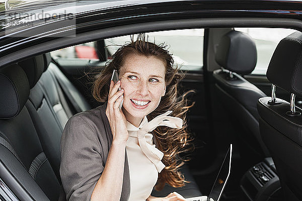 Deutschland  Porträt einer lächelnden Geschäftsfrau im Auto beim Telefonieren mit dem Smartphone