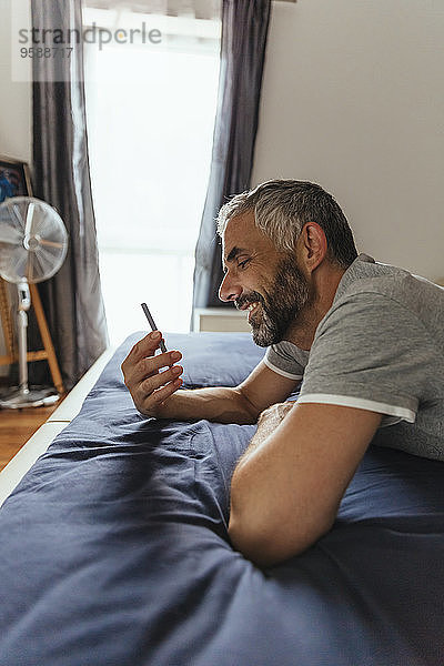 Lächelnder Mann liegt auf seinem Bett und schaut auf sein Smartphone.