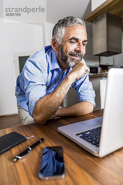 Porträt eines lächelnden Geschäftsmannes im Home-Office mit Blick auf seinen Laptop
