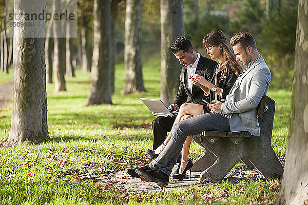 Drei Geschäftsleute sitzen auf einer Parkbank mit Laptop  Smartphone und digitalem Tablett.