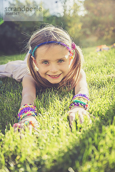 Porträt eines lächelnden kleinen Mädchens mit Webstuhlarmbändern und Haarband auf einer Wiese liegend