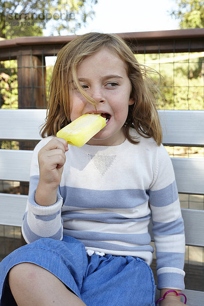 Porträt eines Mädchens  das eisgekühlten Lolly auf der Gartenbank isst.