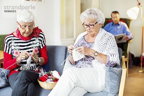 Seniorinnen stricken  während der Mann im Pflegeheim im Hintergrund Buch liest.