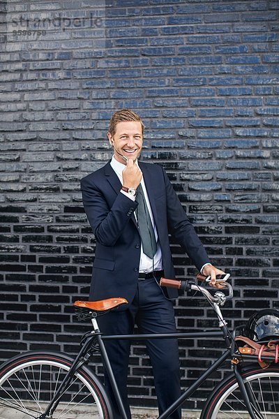 Porträt eines glücklichen Geschäftsmannes  der mit dem Fahrrad auf dem Bürgersteig steht und mit dem Handy telefoniert.