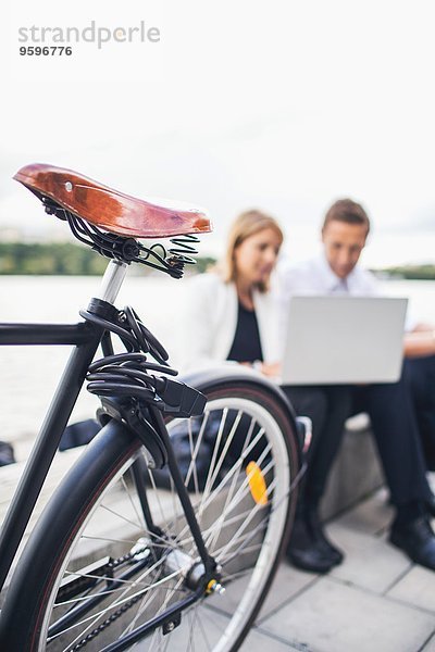Abgeschnittenes Bild des Fahrrads auf dem Bürgersteig mit Geschäftsleuten  die am Laptop gegen den klaren Himmel arbeiten.