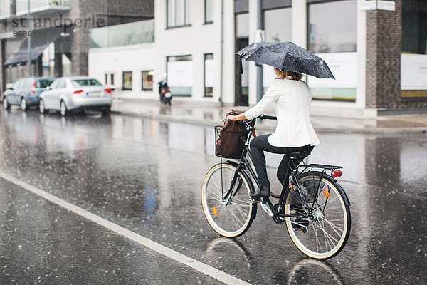 Durchgehende Rückansicht der Geschäftsfrau  die während der Regenzeit auf der nassen Stadtstraße Fahrrad fährt.
