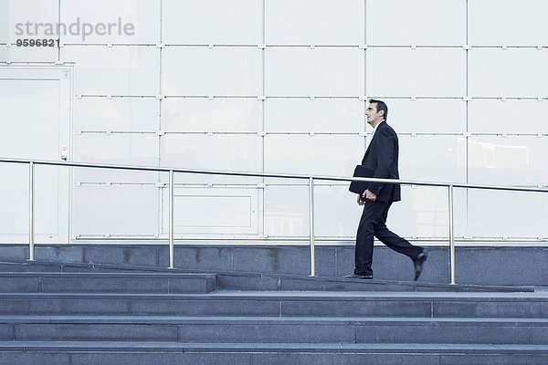 Seitenansicht eines Geschäftsmannes  der an einem Geländer vor dem Bürogebäude entlanggeht.
