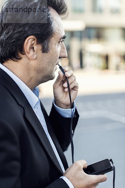 Seitenansicht des Geschäftsmannes  der über Kopfhörer mit dem Handy auf der Straße kommuniziert.