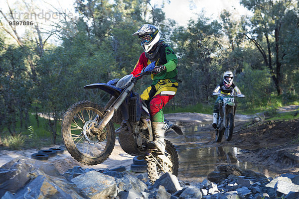 Zwei junge männliche Motocross-Fahrer rasen durch den Waldfluss.