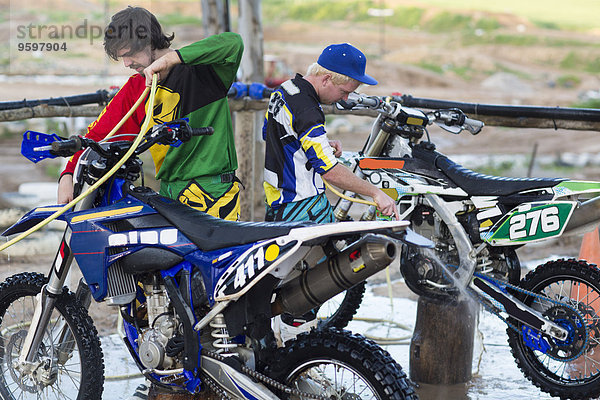 Zwei männliche Motocross-Teilnehmer beim Reinigen von Motorrädern mit Wasserschläuchen