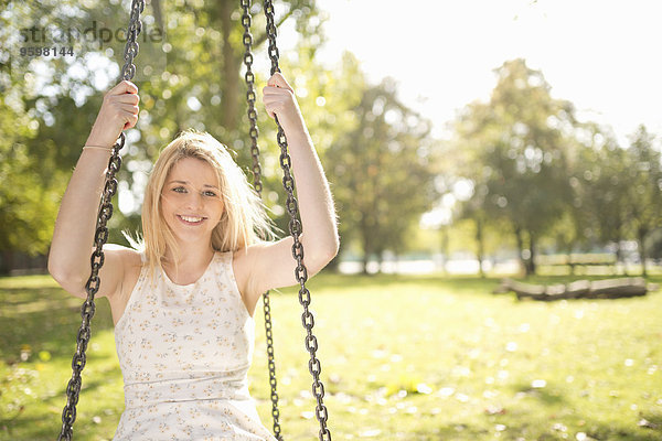 Porträt einer lächelnden jungen Frau  die auf einer Schaukel im Park sitzt.