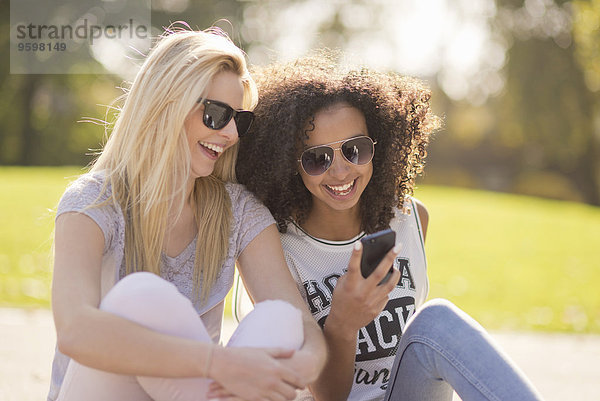 Zwei junge Frauen  die auf eine SMS auf dem Smartphone im Park herabblicken.