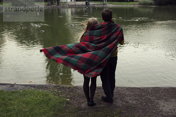 Rückansicht des jungen Paares in Decke gehüllt am Seeufer des Parks