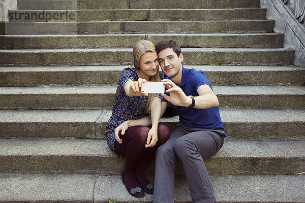 Junges Paar sitzt auf einer alten Strassentreppe und posiert für Smartphone Selfie