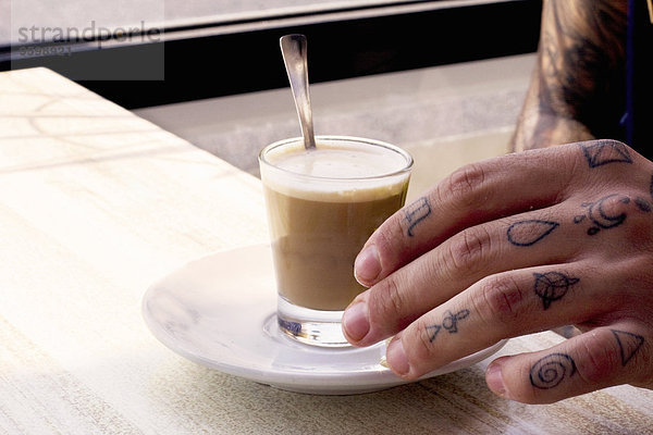 Tätowierte Hand des jungen Mannes und Kaffeeglas auf Kaffeetisch