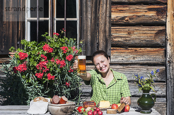 Lächelnde junge Frau sitzt vor der Almhütte und trinkt Bier.