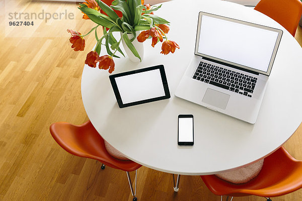 Drei mobile Geräte auf einem weißen runden Tisch mit roten Tulpen und Stühlen