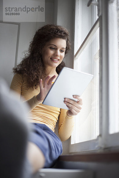 Lächelnde junge Frau mit digitalem Tablett am Fenster