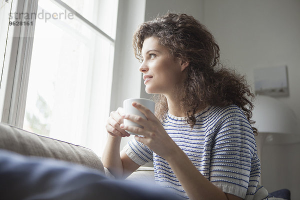 Lächelnde junge Frau mit einer Tasse Kaffee aus dem Fenster schauend