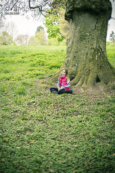 Kleines Mädchen vor einem alten Baum auf einer Wiese sitzend
