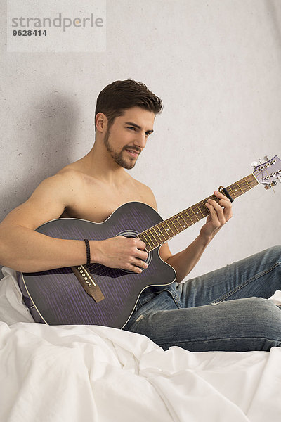 Shirtloser Mann mit Gitarre auf dem Bett sitzend