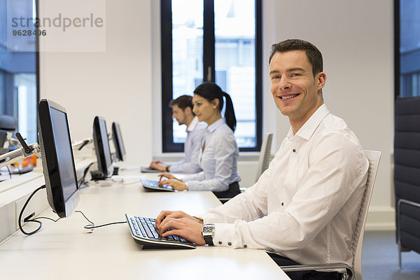Porträt eines lächelnden Mannes am Schreibtisch mit Kollegen im Hintergrund